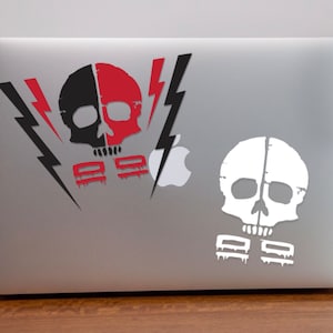 May include: Two skull stickers on a silver laptop. The top sticker is black and red with lightning bolts. The bottom sticker is white with a skull and the text "999".