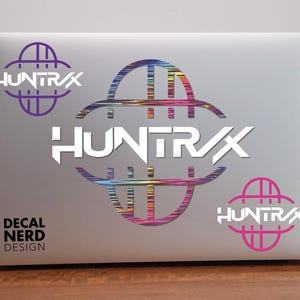 May include: A laptop with a silver finish displays a "HUNTRAX" decal. The design features the word "HUNTRAX" in white, with a stylized graphic in purple, rainbow, and pink. The text "DECAL NERD DESIGN" is also visible.