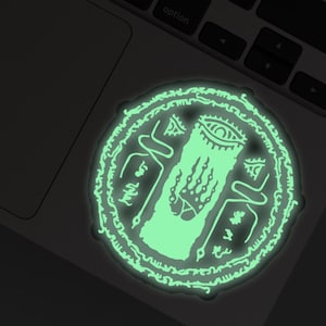 May include: A glowing green sticker featuring a circular design with intricate symbols and an eye at the top. The sticker has a central vertical element and is surrounded by a detailed border. The sticker is placed on a laptop.