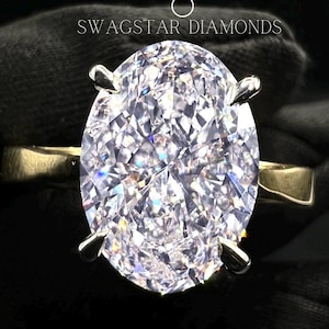 May include: A large, clear oval-shaped diamond ring set in a silver band. The ring is held by a gloved hand. The text "Swagstar Diamonds" is visible in the background.
