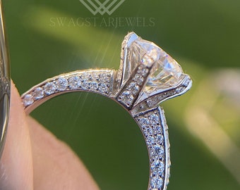 May include: Close-up of a diamond engagement ring. The ring features a large, round-cut diamond set in a prong setting. The band is encrusted with smaller diamonds. The ring is held against a blurred green background. The text "SWAGSTARJEWELS" is visible.