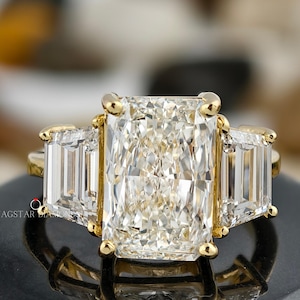 May include: A radiant-cut diamond ring with a gold band. The ring features a large rectangular center stone flanked by two smaller rectangular stones. The ring is set on a black surface, reflecting the light from the diamonds.