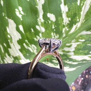 May include: A close-up of a gold and white engagement ring with a large oval-shaped diamond. The ring is set on a black surface with a green leaf in the background.