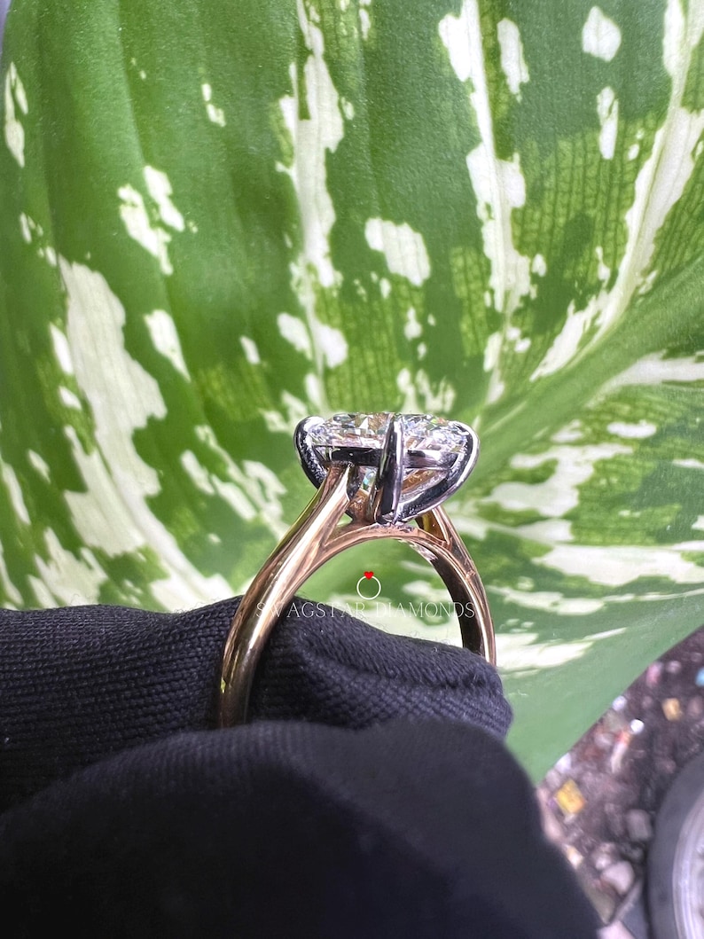 May include: A close-up of a gold and white engagement ring with a large oval-shaped diamond. The ring is set on a black surface with a green leaf in the background.