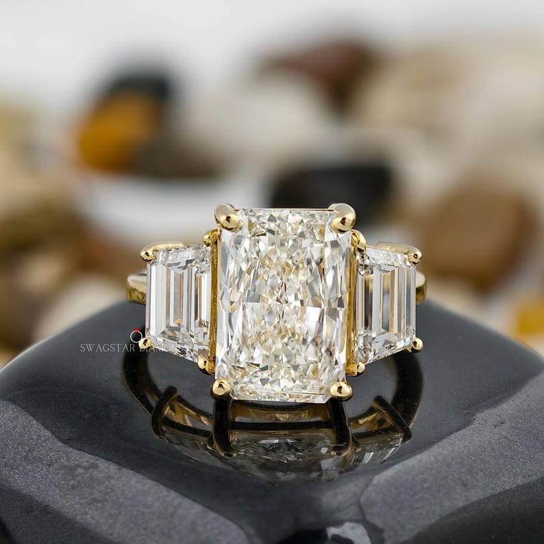 May include: A radiant-cut diamond ring with a gold band. The ring features a large rectangular center stone flanked by two smaller rectangular stones. The ring is set on a black surface, reflecting the light from the diamonds.