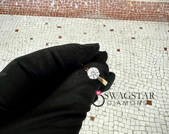 May include: A gold ring featuring a single round brilliant-cut white diamond held in a six-prong setting. The ring is displayed against a black fabric background with the text SWAGSTAR DIAMONDS printed in black near the lower right corner.