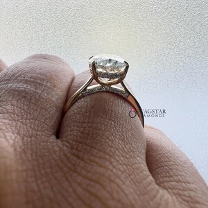 May include: A gold engagement ring with a large, round, clear gemstone. The ring has a delicate band with smaller accent stones. The ring is held up by a hand, showcasing the design and sparkle of the ring.