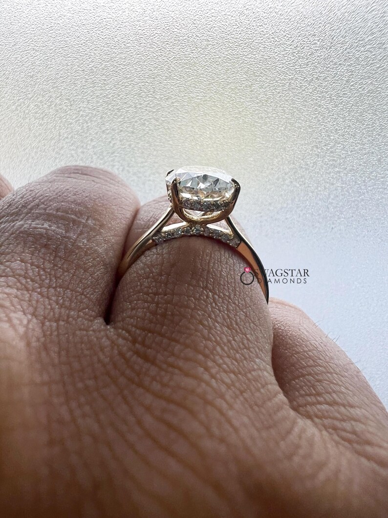 May include: A gold engagement ring with a large, round, clear gemstone. The ring has a delicate band with smaller accent stones. The ring is held up by a hand, showcasing the design and sparkle of the ring.