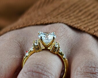 May include: A gold ring featuring a large, round, brilliant-cut clear center stone held in a prong setting, flanked by smaller clear stones set along the band. The text SWAGSTAR DIAMONDS is visible on the side of the gold band.