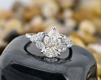May include: A pear-shaped diamond ring with smaller accent diamonds. The ring features a large, central pear-cut diamond and is set in a silver band. The ring is displayed on a black, glossy surface.