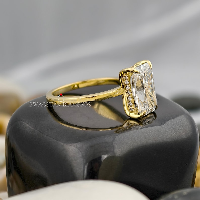 May include: A radiant-cut diamond ring with a gold band. The diamond is set in a gold bezel with smaller diamonds along the sides. The ring is displayed on a black surface. The text "SWAGSTAR DIAMONDS" is visible.