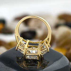 May include: A gold ring with three large, square-cut diamonds. The ring's band and setting are also gold, with a detailed design around the diamonds. The ring is set on a black surface, with a blurred background.