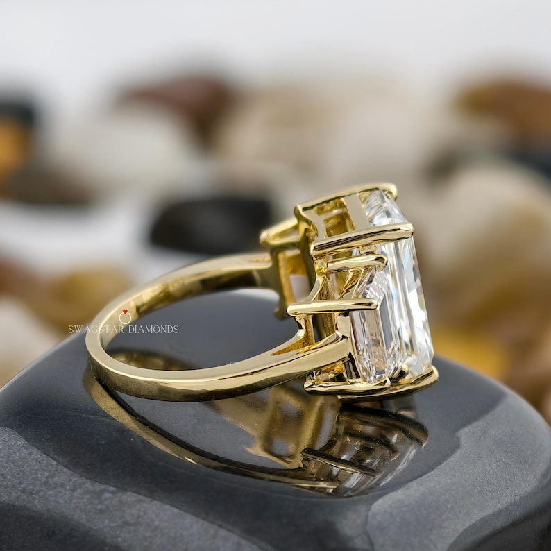 May include: A gold ring with a large, rectangular, clear gemstone as the primary stone, flanked by two smaller, clear, rectangular gemstones. The ring is set on a dark, reflective surface. The brand name "Swag Star Diamonds" is visible on the ring.