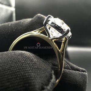 May include: A gold and white engagement ring with a large, round, clear diamond. The ring is set in a prong setting and has a split shank. The ring is on a black velvet background. The text "Swagstar Diamonds" is visible on the ring.