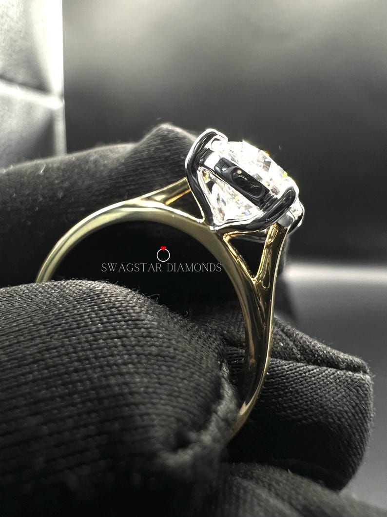 May include: A gold and white engagement ring with a large, round, clear diamond. The ring is set in a prong setting and has a split shank. The ring is on a black velvet background. The text "Swagstar Diamonds" is visible on the ring.