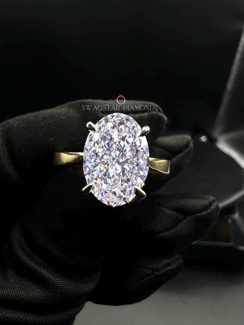May include: A large, clear oval-shaped diamond ring set in a silver band. The ring is held by a gloved hand. The text "Swagstar Diamonds" is visible in the background.