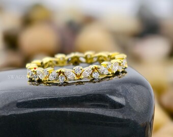 May include: A gold band ring featuring a repeating pattern of marquise-cut and round-cut clear stones set in a continuous row. The metal has a polished yellow finish, and the stones are arranged to create a floral-inspired, symmetrical design.