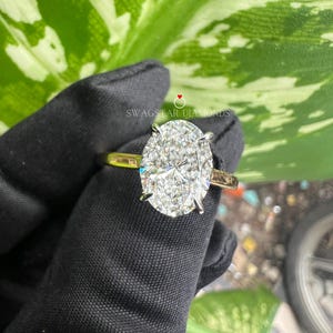 May include: A close-up of a gold engagement ring with a large oval-shaped diamond. The diamond is set in a pave setting and has a brilliant cut. The ring is on a black background.