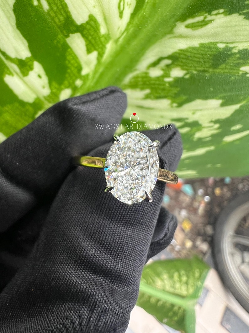 May include: A close-up of a gold engagement ring with a large oval-shaped diamond. The diamond is set in a pave setting and has a brilliant cut. The ring is on a black background.