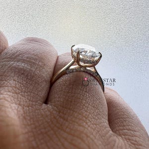 May include: Close-up of a gold ring featuring a large, oval-cut diamond as the centerpiece. The band is also adorned with smaller diamonds. The ring is set against a neutral background, highlighting the sparkle of the gemstones. The ring is a piece of jewelry.