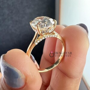 May include: A gold engagement ring with a large, clear, round-cut diamond as the centerpiece. The ring features a halo of smaller diamonds around the main stone and along the band. The ring is held up by a hand with a light blue manicure.