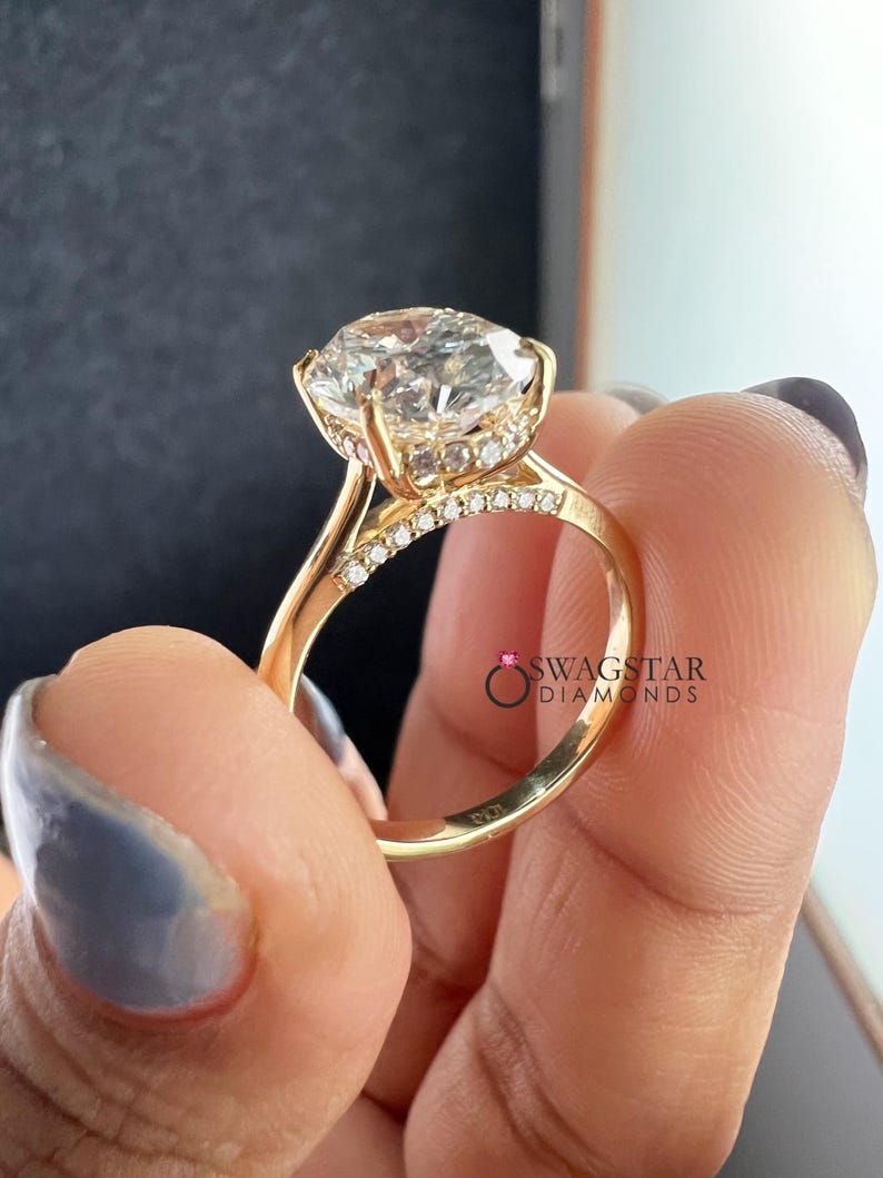 May include: A gold engagement ring with a large, clear, round-cut diamond as the centerpiece. The ring features a halo of smaller diamonds around the main stone and along the band. The ring is held up by a hand with a light blue manicure.