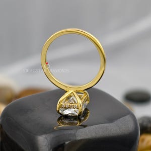 May include: A gold ring with a large, clear, square-cut gemstone and smaller accent stones. The ring band is smooth and polished. The ring is set on a dark, reflective surface. The text "SWAGSTAR DIAMONDS" is visible.