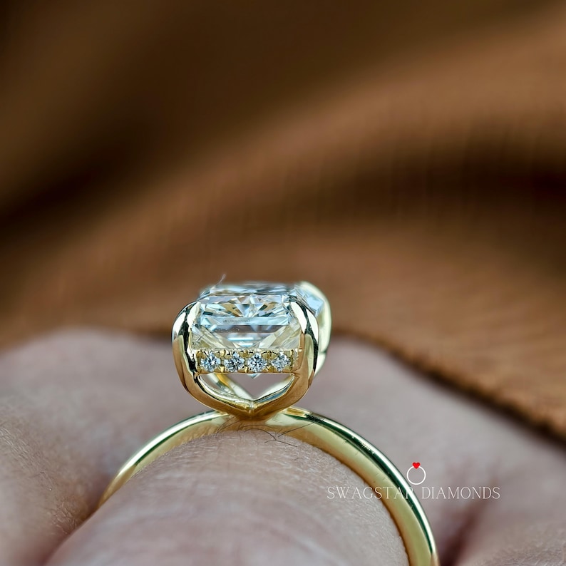 May include: Close-up of a gold ring featuring a large, rectangular-cut diamond. The ring has a delicate band and a setting with smaller accent diamonds. The ring is set against a blurred background.