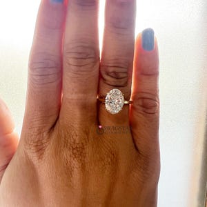 May include: A gold ring with an oval-cut diamond. The ring is on a person's hand with blue nail polish. The diamond is the focal point, reflecting light. The ring's band is thin and simple. The image is well-lit, highlighting the ring's details.