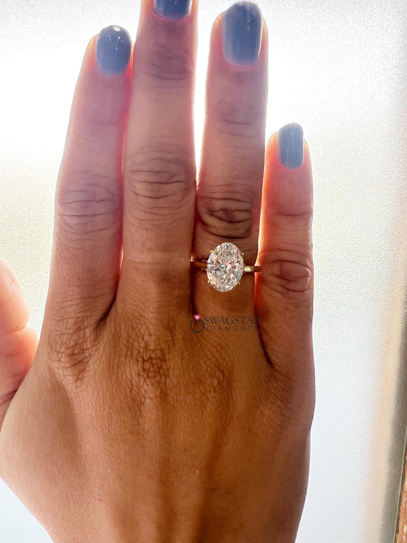 May include: A gold ring with an oval-cut diamond. The ring is on a person's hand with blue nail polish. The diamond is the focal point, reflecting light. The ring's band is thin and simple. The image is well-lit, highlighting the ring's details.
