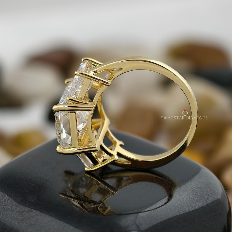 May include: A gold ring with three large, clear, rectangular-cut stones. The ring's band and setting are a bright gold color. The ring is displayed on a black surface. The text "SWAGSTAR DIAMONDS" is visible on the ring.