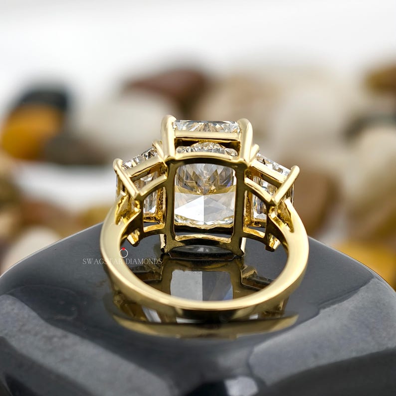 May include: A gold ring with a large, rectangular, clear gemstone as the center stone, flanked by two smaller, rectangular gemstones on each side. The ring is set on a black surface. The brand name "SWAG OF DIAMONDS" is visible.