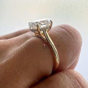 May include: A gold engagement ring featuring a large, oval-cut diamond. The ring has a delicate band and a halo of smaller diamonds around the main stone. The ring is shown from a side angle.