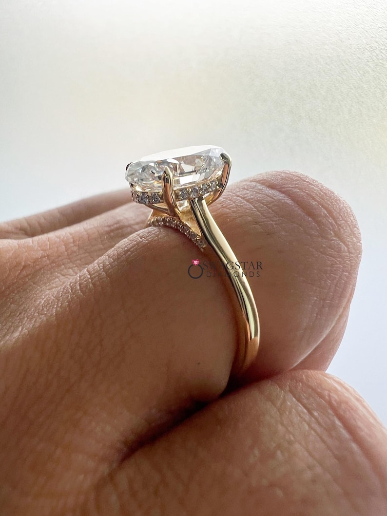 May include: A gold engagement ring featuring a large, oval-cut diamond. The ring has a delicate band and a halo of smaller diamonds around the main stone. The ring is shown from a side angle.