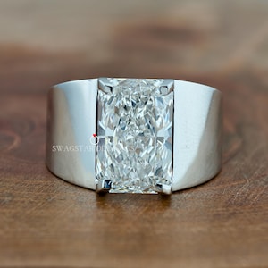 May include: A silver ring with a large, rectangular, clear gemstone. The ring has a wide band and a modern design. The gemstone is the focal point, catching the light and reflecting a brilliant sparkle. The ring is set on a wooden surface.