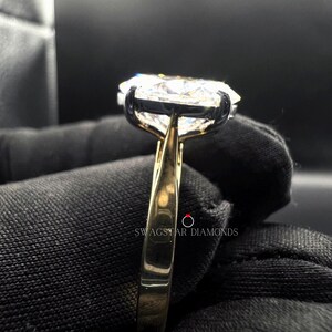May include: A gold band engagement ring with a large, clear, round diamond set in a prong setting. The ring is on a black fabric background.