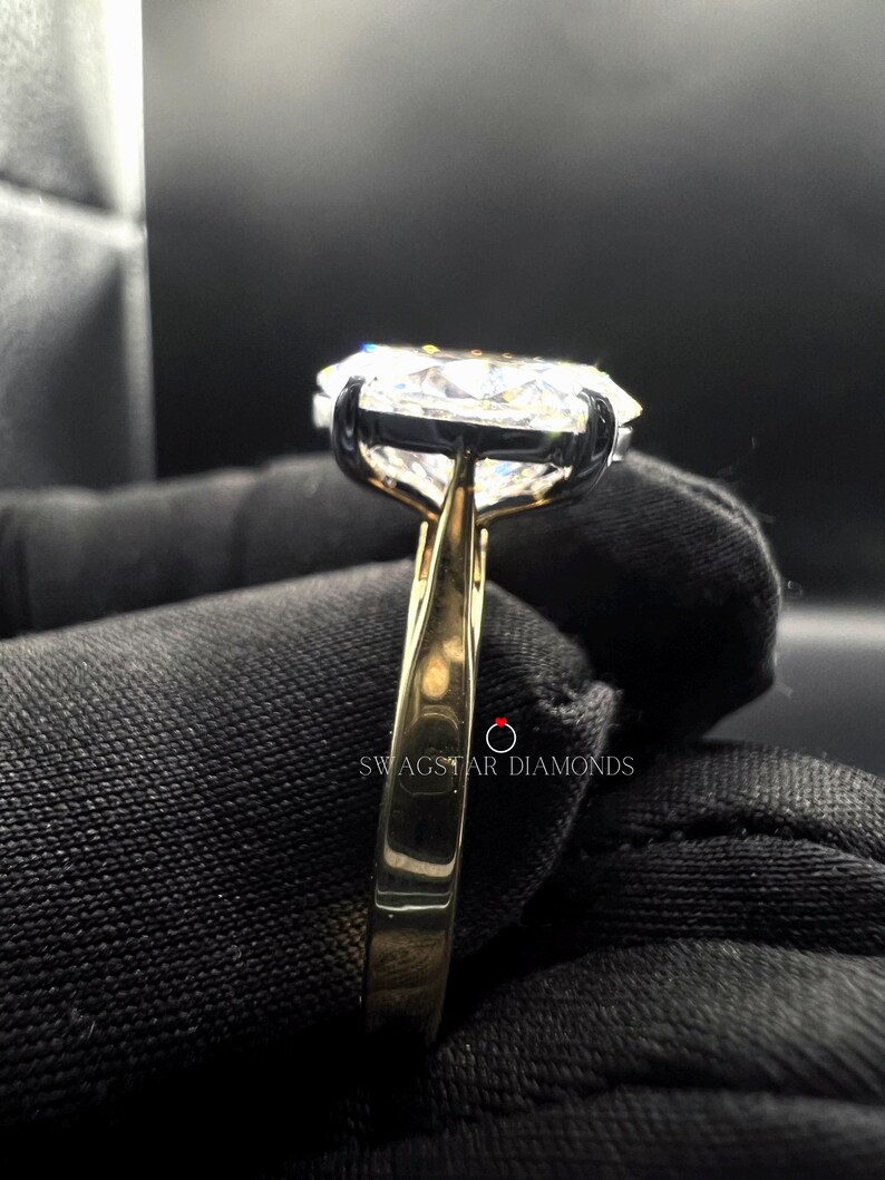 May include: A gold band engagement ring with a large, clear, round diamond set in a prong setting. The ring is on a black fabric background.