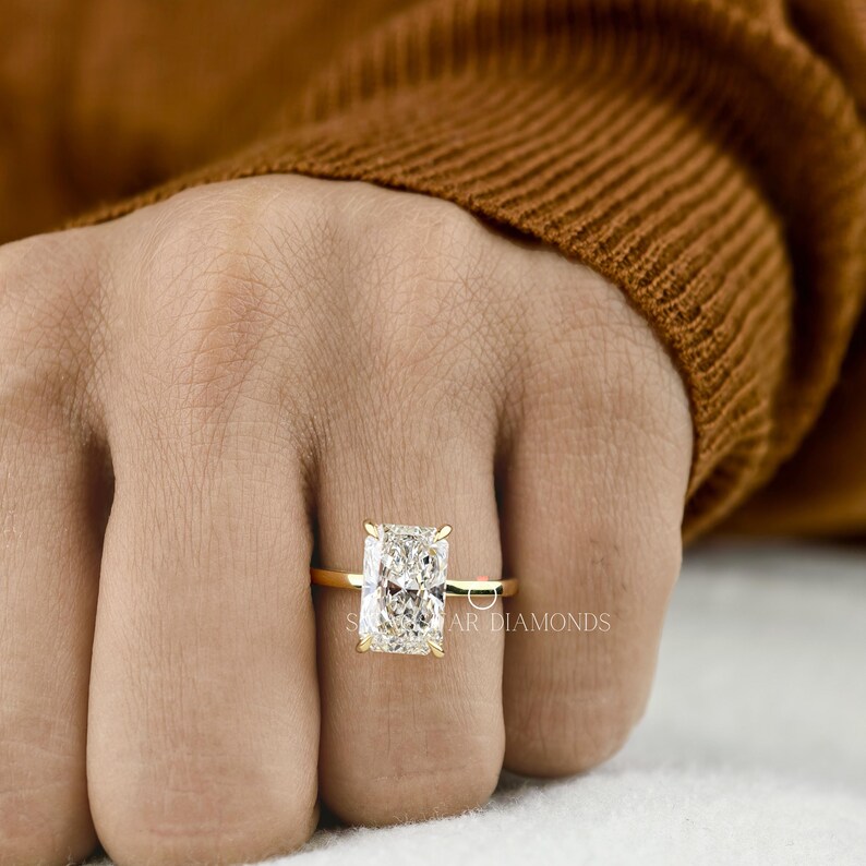 May include: A gold ring with a large, rectangular, clear diamond. The ring is set on a gold band. The diamond is the focal point of the ring, reflecting light. The ring is on a hand.