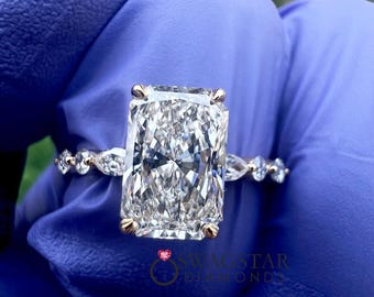 May include: A radiant-cut diamond ring featuring a large rectangular center stone held by four prongs on a thin band. The band is accented with smaller marquise and round-cut diamonds on each side. The text SWAGSTAR DIAMONDS is visible.