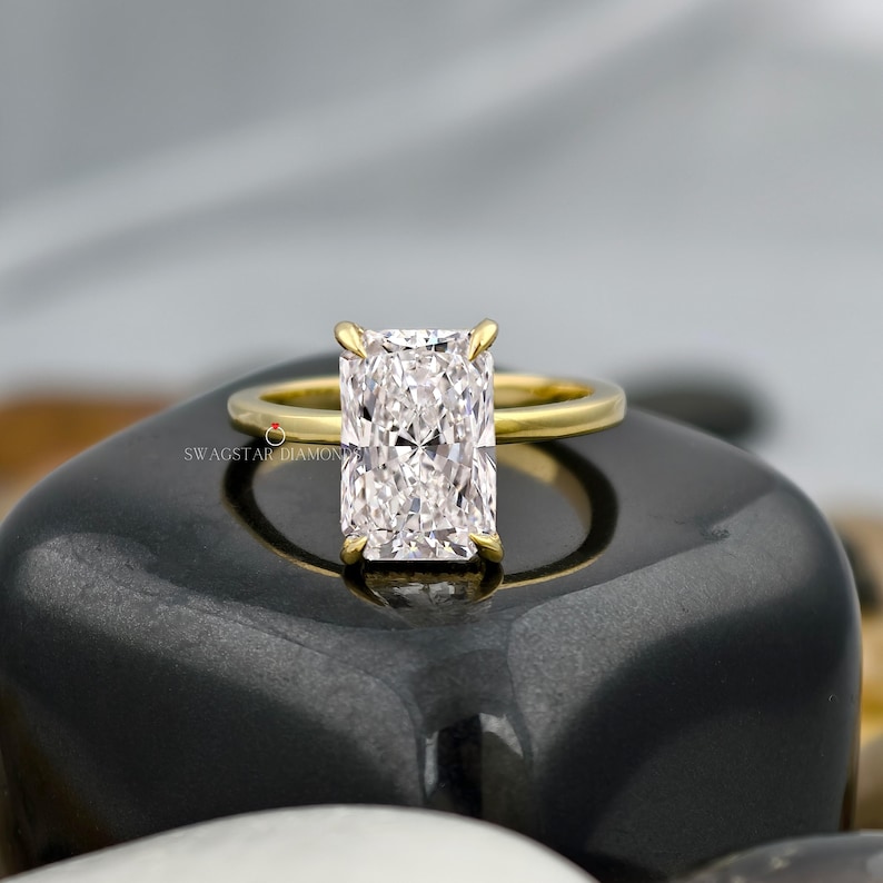 May include: A radiant-cut diamond ring with a gold band. The rectangular diamond is set in a four-prong setting. The ring is displayed on a black, glossy surface. The ring is a piece of jewelry.