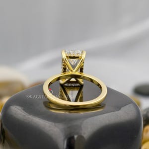 May include: A gold engagement ring with a large, square-cut diamond. The ring's band and setting are gold, and the diamond is clear and sparkling. The ring is displayed on a dark, reflective surface.