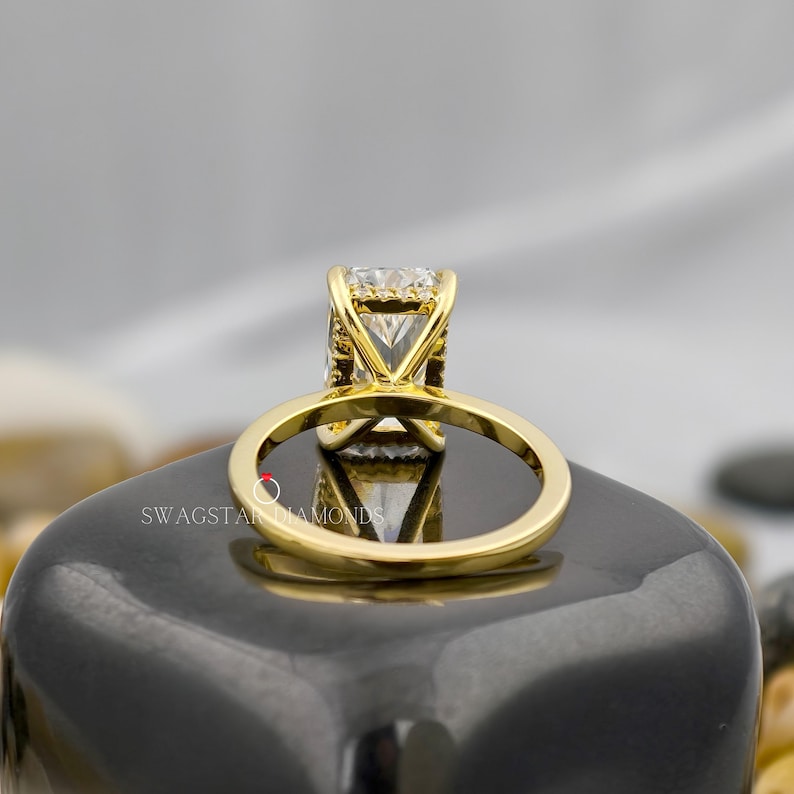 May include: A gold engagement ring with a large, square-cut diamond. The ring's band and setting are gold, and the diamond is clear and sparkling. The ring is displayed on a dark, reflective surface.