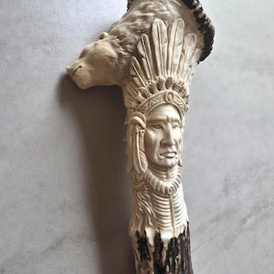 Peut inclure: Une sculpture en corne sculptée représentant un chef amérindien avec une coiffe de plumes et une tête d'ours. La sculpture est principalement blanc cassé avec des accents brun foncé à la base et au sommet.