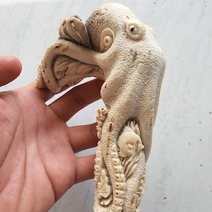 Op de afbeelding: Een gesneden ivoorkleurig octopusbeeldje met gedetailleerde tentakels en een getextureerd oppervlak. De octopus is gebeeldhouwd met een vis en ander zeeleven. De basis is donkerbruin.