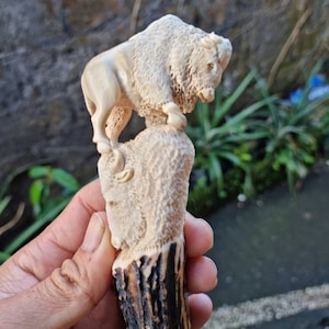 K&ouml;nnte beinhalten: Ein handgeschnitzter Flaschenverschluss aus Holz mit einer detaillierten Bison-Skulptur. Der Bison ist in hellbrauner Farbe geschnitzt und steht auf einem strukturierten Sockel. Der Sockel ist an einem dunkelbraunen Hirschhorn-Griff befestigt.