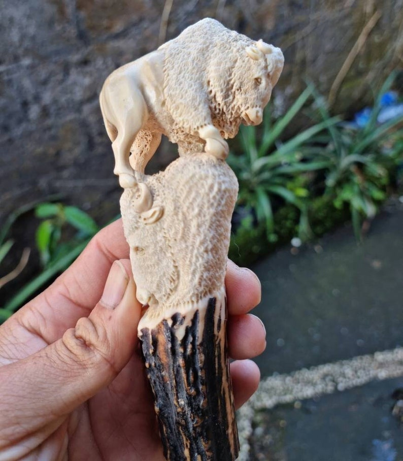 K&ouml;nnte beinhalten: Ein handgeschnitzter Flaschenverschluss aus Holz mit einer detaillierten Bison-Skulptur. Der Bison ist in hellbrauner Farbe geschnitzt und steht auf einem strukturierten Sockel. Der Sockel ist an einem dunkelbraunen Hirschhorn-Griff befestigt.