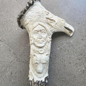 May include: A carved antler sculpture depicting a Native American face, an eagle head, and a bear's face. The off-white carving has a dark brown, textured base. The eagle's beak is open, and the Native American figure wears a headdress.