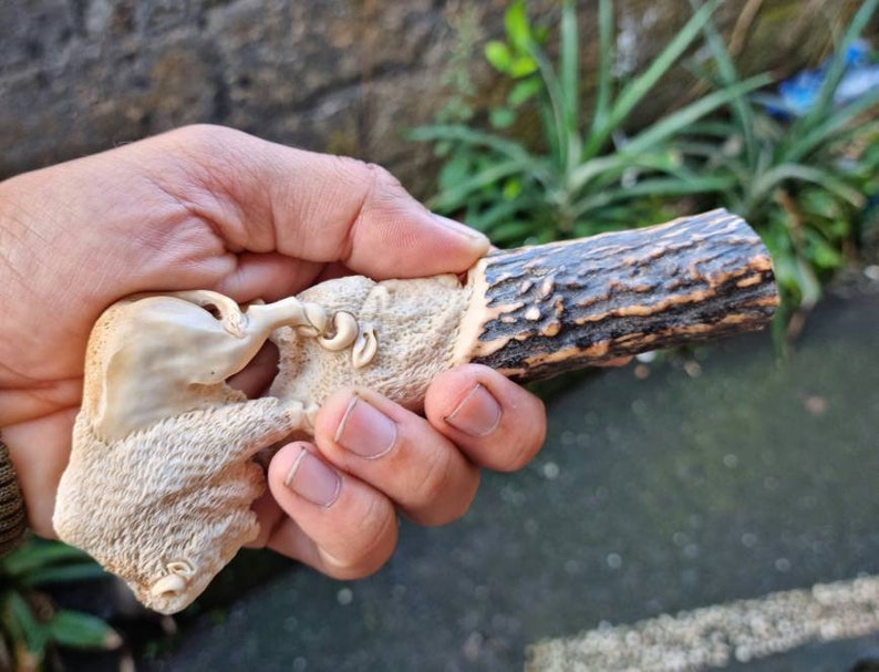 K&ouml;nnte beinhalten: Eine handgeschnitzte Holzskulptur von zwei Bisons, einer steht auf dem anderen. Die Skulptur ist aus hellem braunem Holz gefertigt und hat eine nat&uuml;rliche Oberfl&auml;che. Die Skulptur ist auf einem St&uuml;ck Geweih montiert.