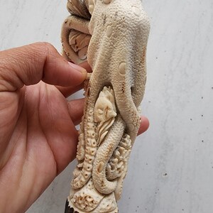 May include: A detailed, off-white carved sculpture of an octopus with a fish and other sea creatures. The sculpture is held in a hand, showcasing the intricate details and textures of the artwork. The base appears to be made of a dark brown material.