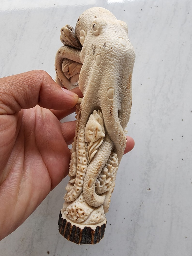 May include: A detailed, off-white carved sculpture of an octopus with a fish and other sea creatures. The sculpture is held in a hand, showcasing the intricate details and textures of the artwork. The base appears to be made of a dark brown material.